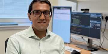 A man with short dark hair and glasses, wearing a light grey shirt, sits at a desk facing the camera.  Two screens and a tablet are visible on the desk behind him.